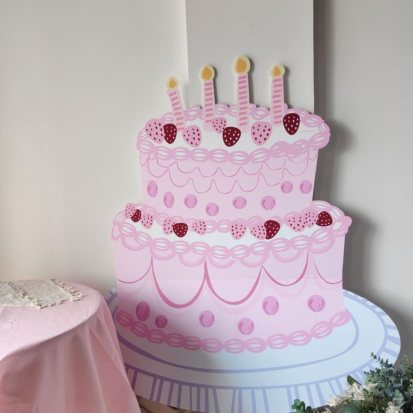 A large, decorative, two-tiered cardboard birthday cake standee cutout stands next to a table draped in a pink tablecloth. The 5ft Happy Birthday Cake Foam Board Cutcout Props Standee by Dongguan Quanjia Trade Co.,ltd is pink with strawberry decorations, swirls, and four unlit candles on top. The table has text on it and some green foliage is visible at the bottom right.