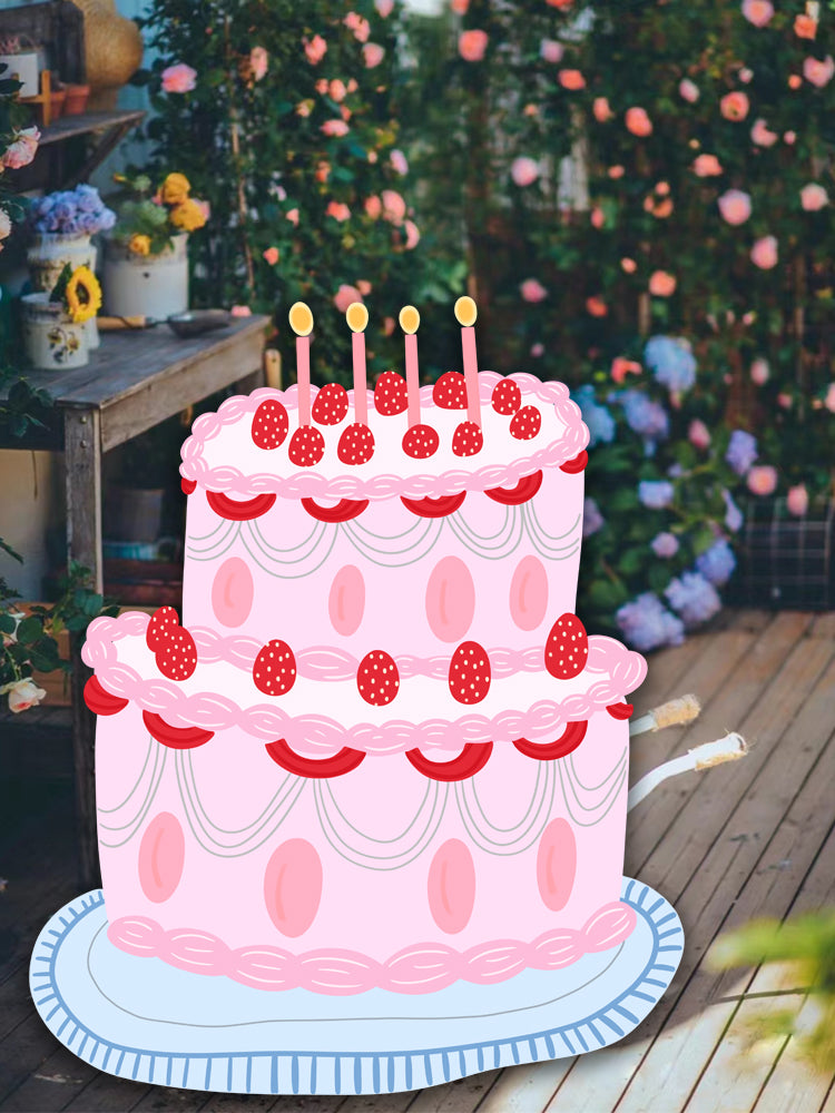 A two-tiered cartoon cake with pink icing, decorated with strawberries, and six unlit candles on top. The cake sits on a patterned plate beside Dongguan Quanjia Trade Co.,ltd 5ft Happy Birthday Cake Foam Board Cutout Props Standee for the party. The background features a wooden garden setting with blooming flowers and plants.