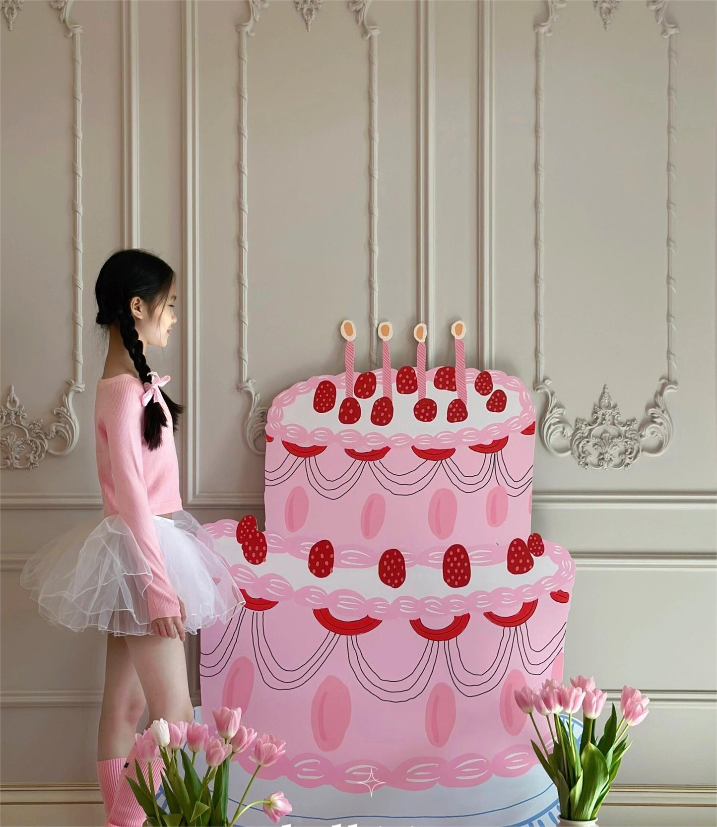 A young girl in a pink top and white tulle skirt stands beside a large, two-tiered pink cake with strawberries and candles. The room is elegantly decorated with ornate wall details, a floor vase filled with pink flowers, and the Dongguan Quanjia Trade Co.,ltd 5ft Happy Birthday Cake Foam Board Cutout Props Standee adding an extra touch of charm to the party.