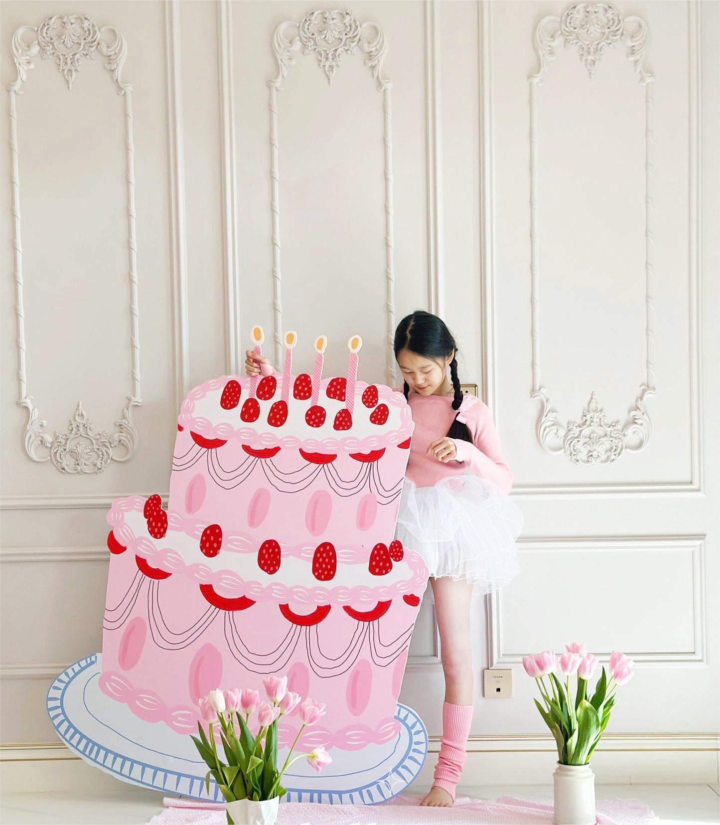 A young girl in a ballet outfit stands indoors holding a large, illustrated, pink three-tiered 5ft Happy Birthday Cake Foam Board Cutcout Props Standee by Dongguan Quanjia Trade Co.,ltd with red strawberries and candles. The room has ornate white paneling on the walls and a vase of pink tulips on the floor in front of her, resembling charming standee cutouts for party decor.