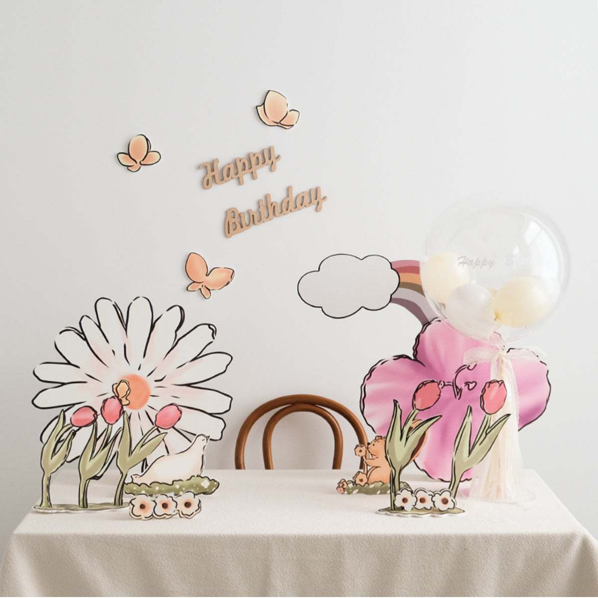 A birthday party setup with a whimsical floral theme. The table features large paper flowers, Eanjia Rainbow Tulip Garden Cutouts | 16pc Watercolor Daisy & Squirrel Decor | Baby Shower & Birthday Props decorations, and a clear balloon with "Happy" written on it. "Happy birthday" and butterfly standee cutouts decorate the white wall behind the table.