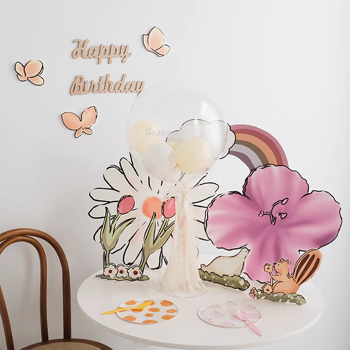 A cheerful birthday setup featuring pastel decorations. A "Happy Birthday" sign with butterflies is on the wall. A table adorned with a large balloon, flower cutouts, a rainbow, and Eanjia's Rainbow Tulip Garden Cutouts | 16pc Watercolor Daisy & Squirrel Decor | Baby Shower & Birthday Props has plates and utensils. A wooden chair stands beside the table.