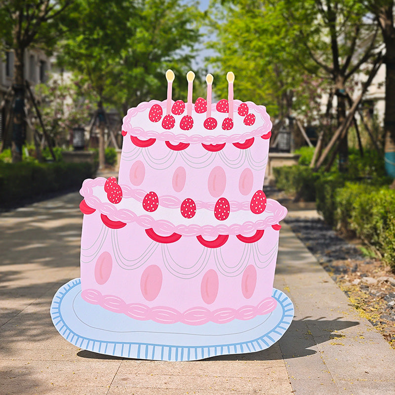 A large, two-tiered pink cartoon "5ft Happy Birthday Cake Foam Board Cutout Props Standee" by Dongguan Quanjia Trade Co.,ltd with red strawberries and lit candles sits on a blue platter. The standee is outdoors on a paved path, surrounded by other standee cutouts for the party, with green trees and bushes lining the sides, and buildings visible in the background.