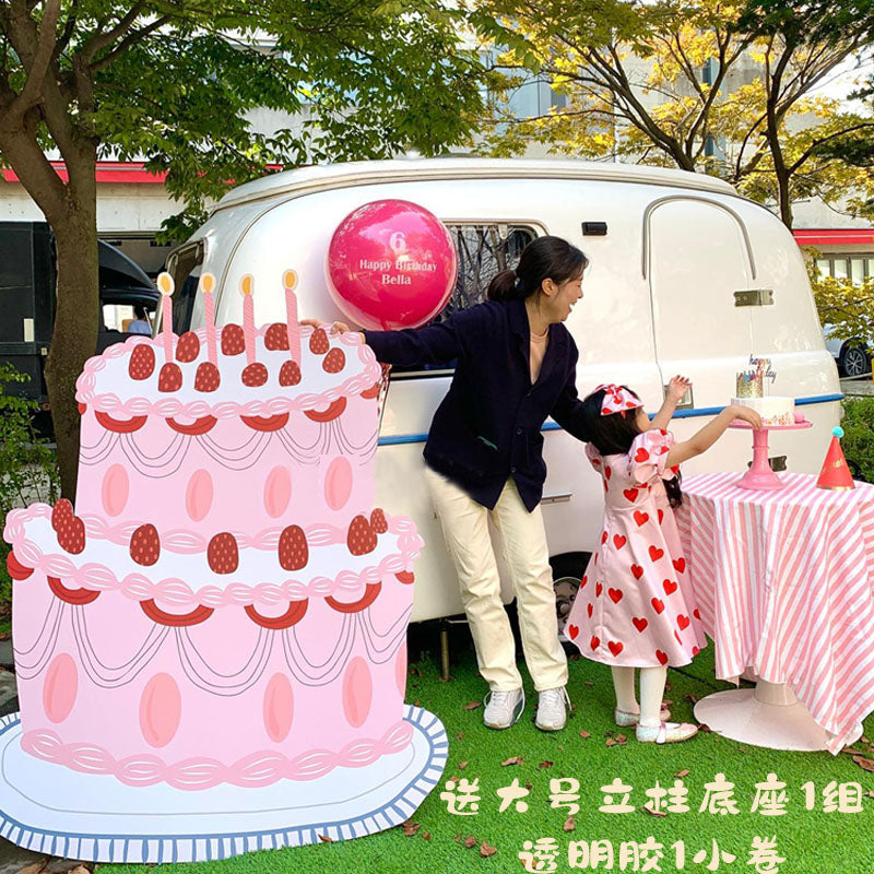 A woman and a child are standing outside near a white trailer decorated for a celebration, surrounded by festive standee cutouts for the party. They are in front of a large, pink, two-tiered 5ft Happy Birthday Cake Foam Board Cutout Props Standee by Dongguan Quanjia Trade Co.,ltd. The child, dressed in a red and white polka dot dress, reaches across a small table with a striped tablecloth.