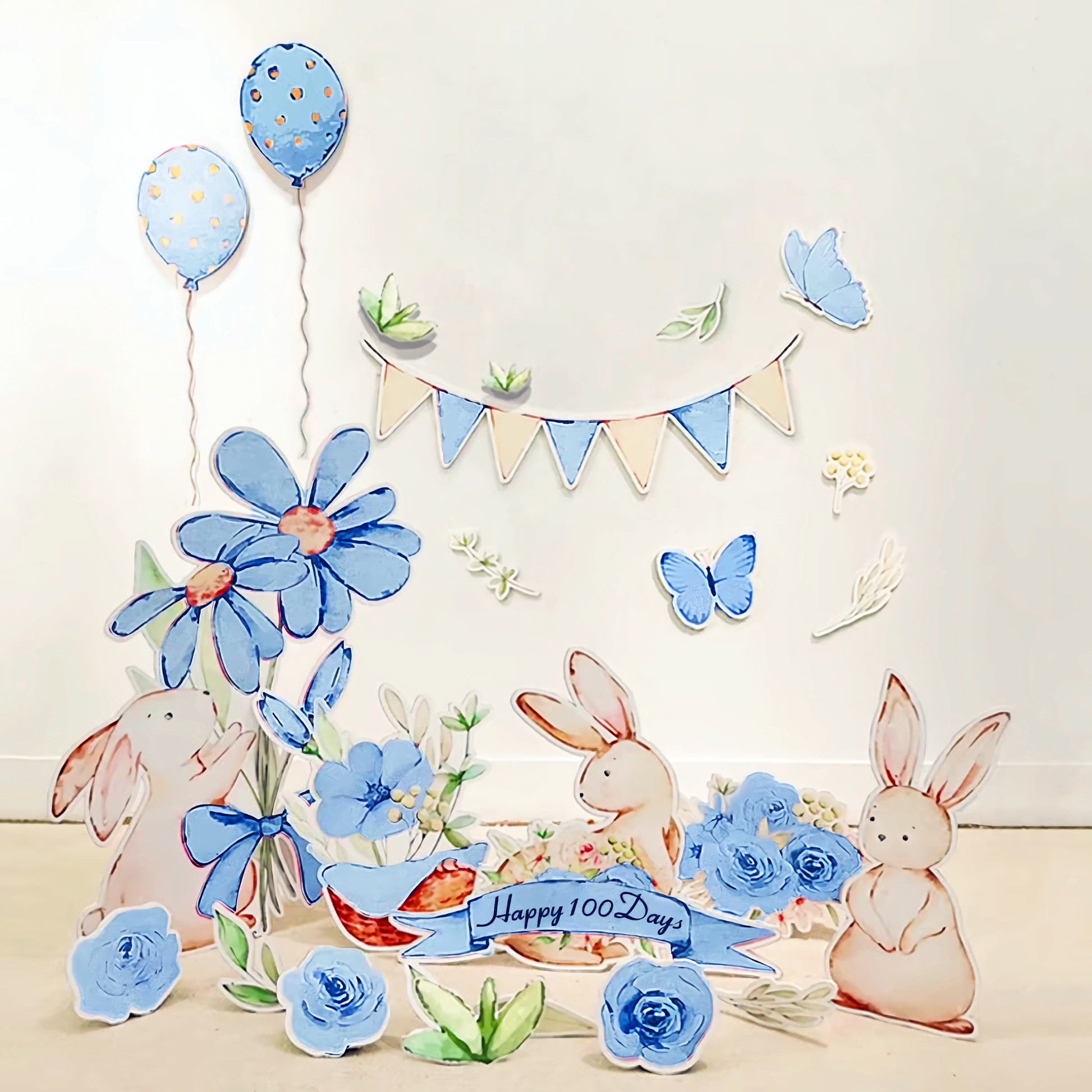 A whimsical scene featuring paper cutouts of rabbits, blue flowers, and butterflies. "Happy 100 Days" is written on a banner. The backdrop includes two blue balloons with gold dots and a pastel bunting banner hanging above, with the Blue Garden Bunny Flower Standee Cut Out Foam Board Decoration Set of 30 from Dongguan Quanjia Trade Co.,ltd completing the charming setup.