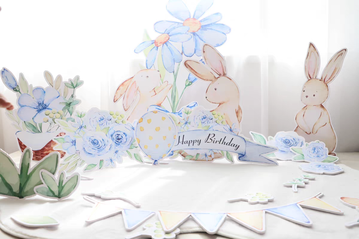 A whimsical birthday setup features the Blue Garden Bunny Flower Standee Cut Out Foam Board Decoration Set of 30 by Dongguan Quanjia Trade Co.,ltd. A "Happy Birthday" sign is prominently displayed amongst blue and yellow floral decorations. Soft light filters through the background, creating a cheerful ambiance.