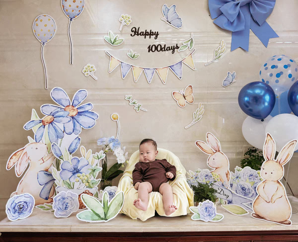 A baby sits in a yellow cushioned chair, surrounded by colorful Blue Garden Bunny Flower Standee Cut Out Foam Board Decoration Set of 30 by Dongguan Quanjia Trade Co.,ltd. Behind the baby are balloons and a festive banner reading "Happy 100days." The setting is bright and cheerful, celebrating the baby's milestone.