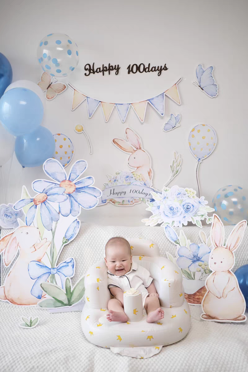 A baby sits in a white cushioned seat, surrounded by blue and white floral and bunny-themed decorations. Balloons, the Blue Garden Bunny Flower Standee Cut Out Foam Board Decoration Set of 30 from Dongguan Quanjia Trade Co.,ltd for the party, and a banner that says "Happy 100days" adorn the background, celebrating the baby's milestone. The scene is cheerful and brightly lit.