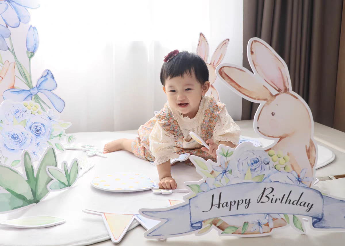 A smiling toddler wearing a floral dress is crawling on a decorated floor amidst pastel-colored birthday decorations. The scene includes the Dongguan Quanjia Trade Co.,ltd Blue Garden Bunny Flower Standee Cut Out Foam Board Decoration Set of 30, featuring standee cutouts of flowers and a bunny holding a "Happy Birthday" banner. White curtains and a brown curtain frame the background.