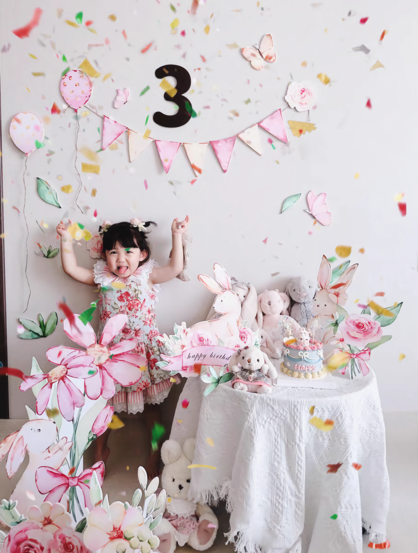 The image shows a joyful toddler wearing a floral dress celebrating her third birthday. She stands beside a decorated table with pastel-colored stuffed animals, flowers, and a small cake. A sign that reads "Happy Birthday" is flanked by Dongguan Quanjia Trade Co.,ltd Pink Bunny Flower Balloons Standee Party Decoration Foam Board Cutout Set 30pcs, while a "3" balloon hangs on the wall and confetti floats in the air.