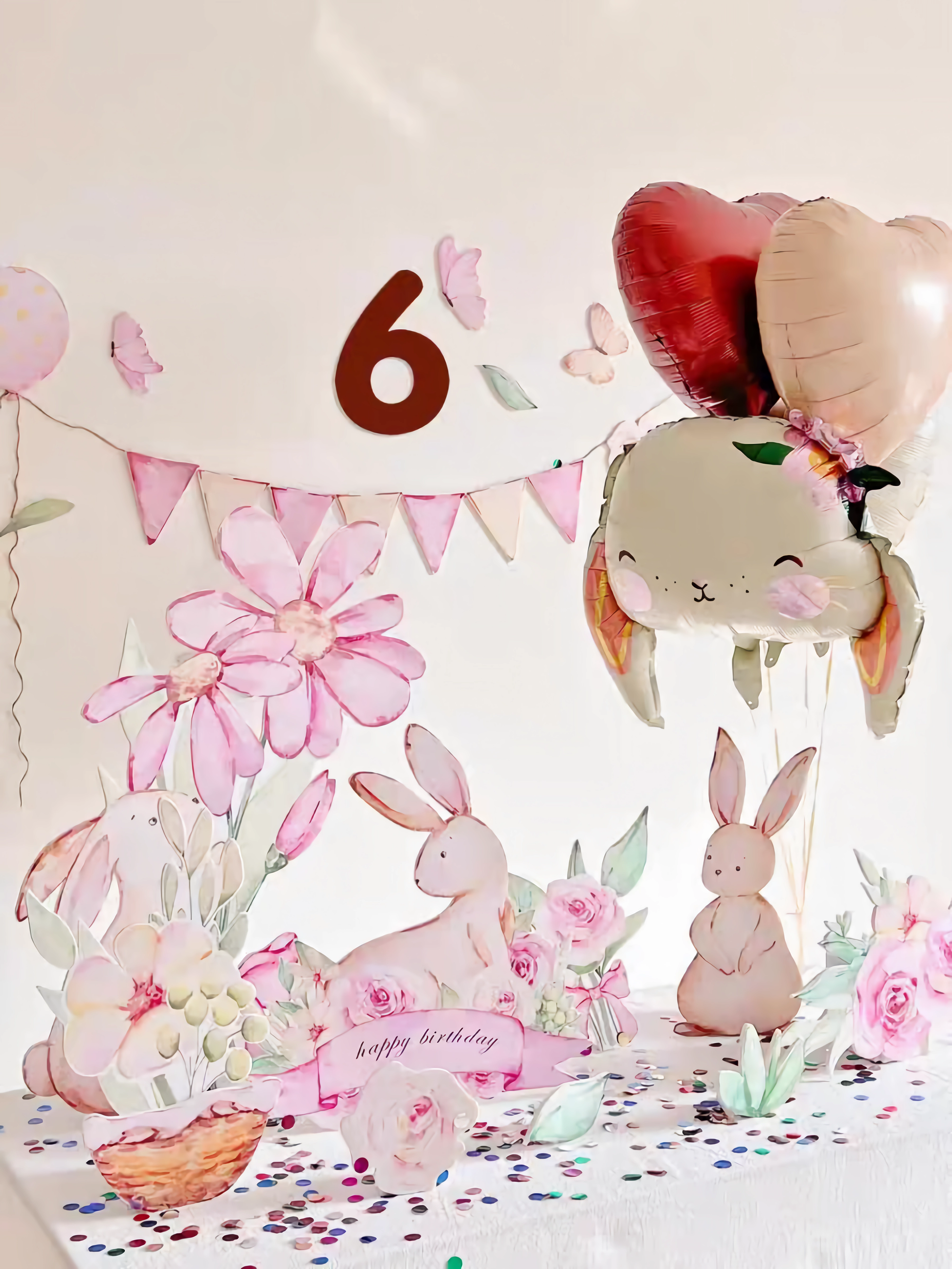 A whimsical birthday setup features a large number six, pink floral decorations, and bunny standee cutouts for the party. Heart-shaped balloons with a bunny face float above the confetti-strewn table. A banner with pink pennants and assorted pastel decorations adorns the wall, completing the festive scene. The entire arrangement is beautifully accentuated by the Pink Bunny Flower Balloons Standee Party Decoration Foam Board Cutout Set 30pcs from Dongguan Quanjia Trade Co.,ltd.